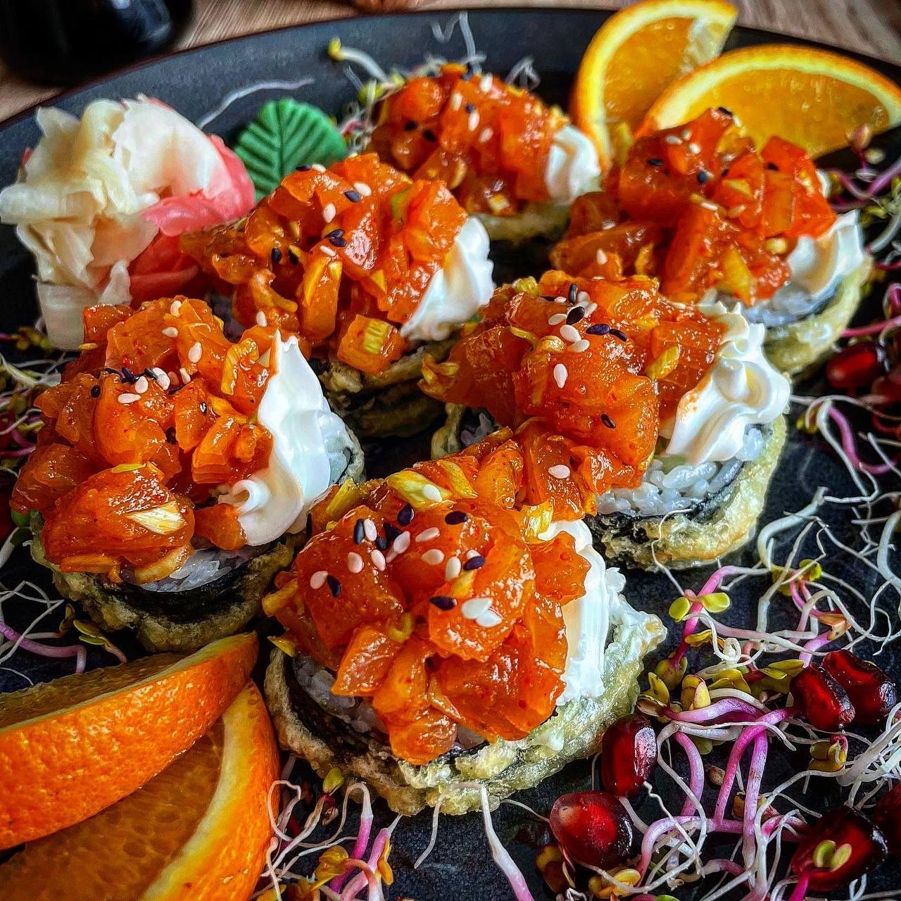 Sześć rolek sushi futomaki w tempurze, z tatarem z łososia, serkiem Philadelphia, posypane sezamem, podane z imbirem, wasabi, kiełkami i pomarańczą na czarnym talerzu.