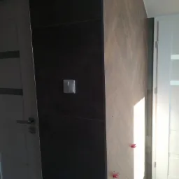 Ściana z ciemnych płytek z włącznikiem światła sąsiaduje z jasną ścianą wyłożoną płytkami w jodełkę, widoczne czerwone krzyżyki dystansowe na płytkach, białe drzwi po lewej i prawej.