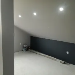 Poddasze w trakcie malowania: ściana pomalowana na ciemny grafit, widoczne puszki z farbą, listwy przypodłogowe i gniazdko elektryczne.