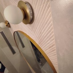 KONSTRUKTOR WM SPÓŁKA Z OGRANICZONĄ ODPOWIEDZIALNOŚCIĄ - Owalne lustro w złotej ramie odbija fragment łazienki z dekoracyjną, beżową ścianą w jodełkę i nowoczesnym oświetleniem w postaci dwóch kulistych lamp.