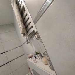 KONSTRUKTOR WM SPÓŁKA Z OGRANICZONĄ ODPOWIEDZIALNOŚCIĄ - Jasna łazienka po remoncie: białe płytki z metalowymi listwami, grzejnik drabinkowy z ręcznikiem, dekoracje na półce. Ukończona aranżacja z dbałością o detale.