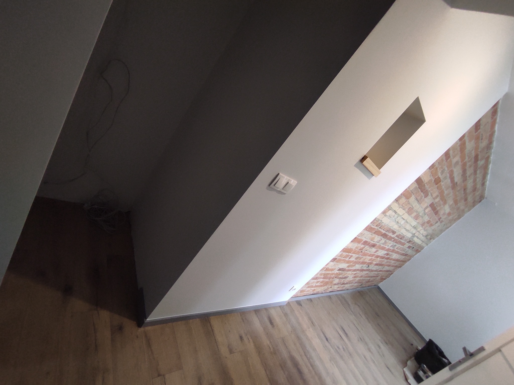 Poddasze po remoncie: ciemnoszara ściana kontrastuje z białą, fragment odsłoniętej cegły, drewniana podłoga, widoczne gniazdko elektryczne i wnęka w ścianie.