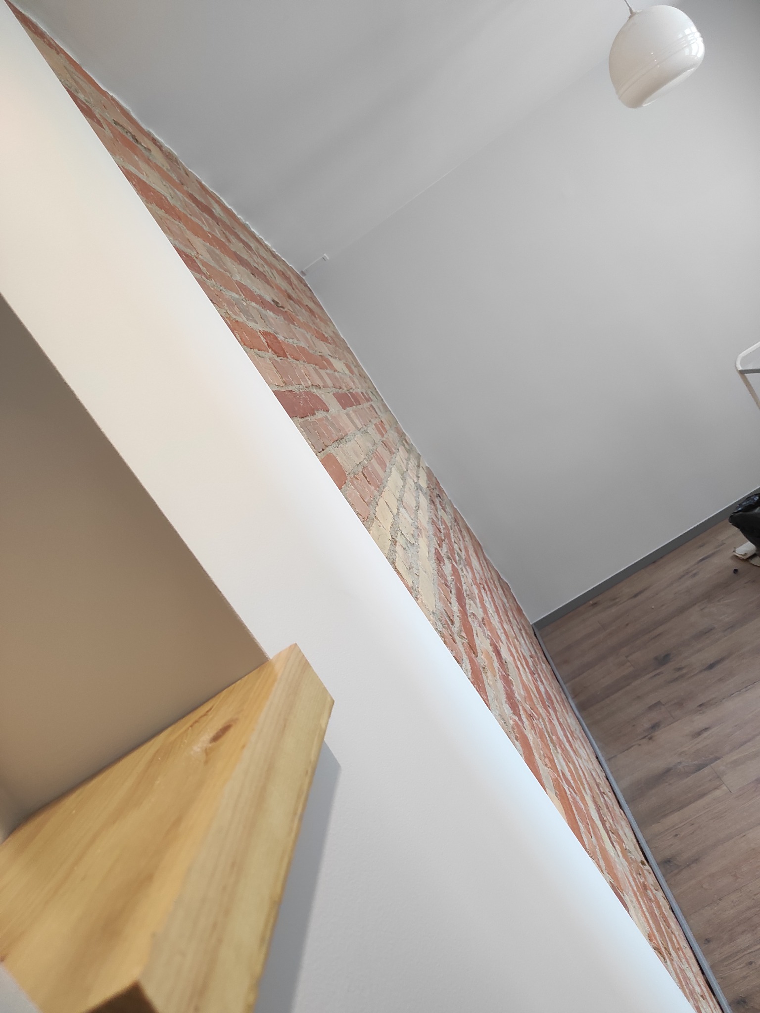 Fragment wnętrza z odsłoniętą cegłą, pomalowaną białą ścianą, drewnianą półką i lampą sufitową, uchwycony pod kątem.