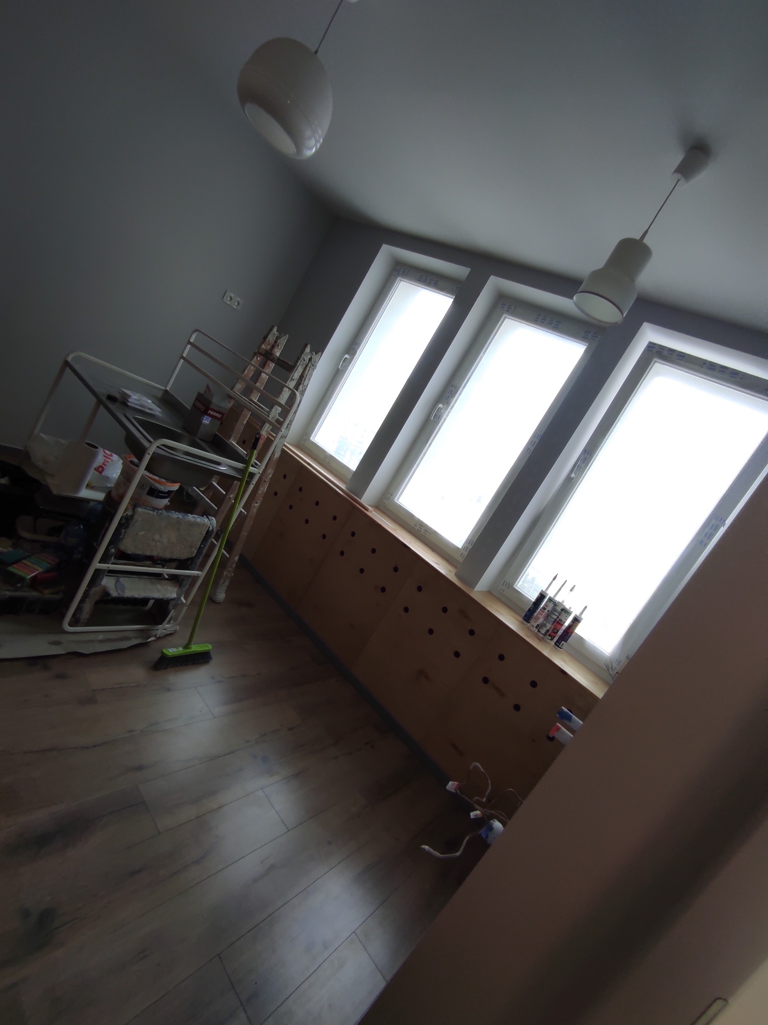 Pokój w trakcie remontu: widoczne trzy okna dachowe, drewniana zabudowa pod oknami z rzędami otworów, drabina, wózek z narzędziami malarskimi, mop, podłoga z paneli.