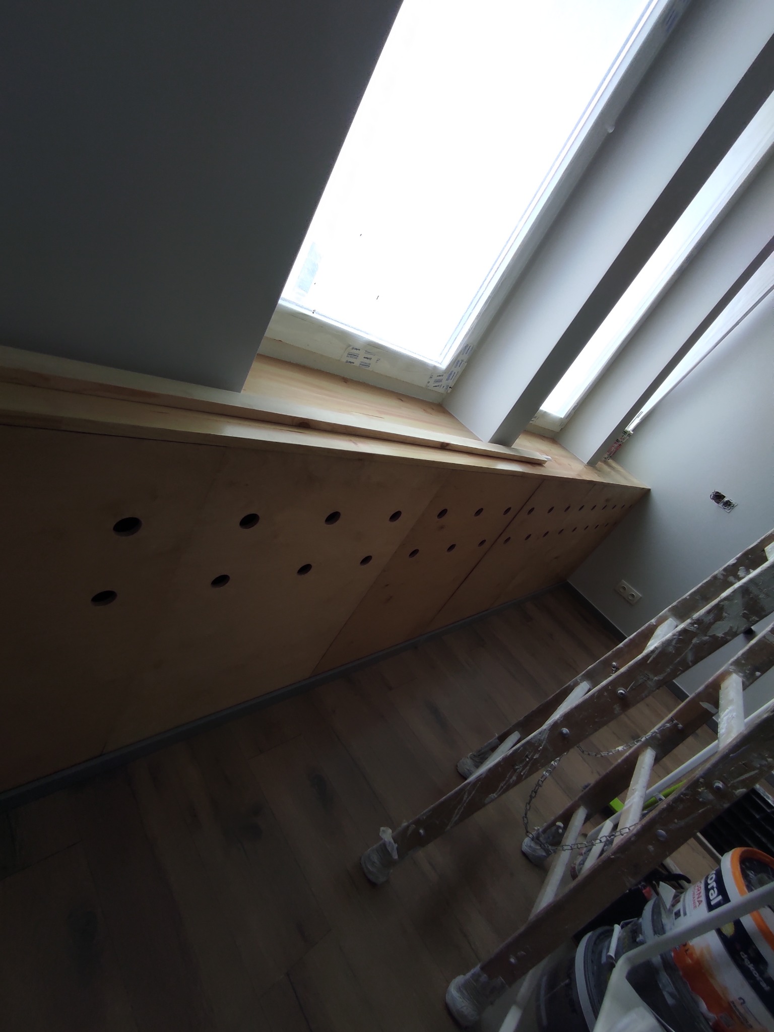 Montaż drewnianej zabudowy pod skosem okna dachowego z wywierconymi otworami, widoczna drabina malarska i puszki z farbą.