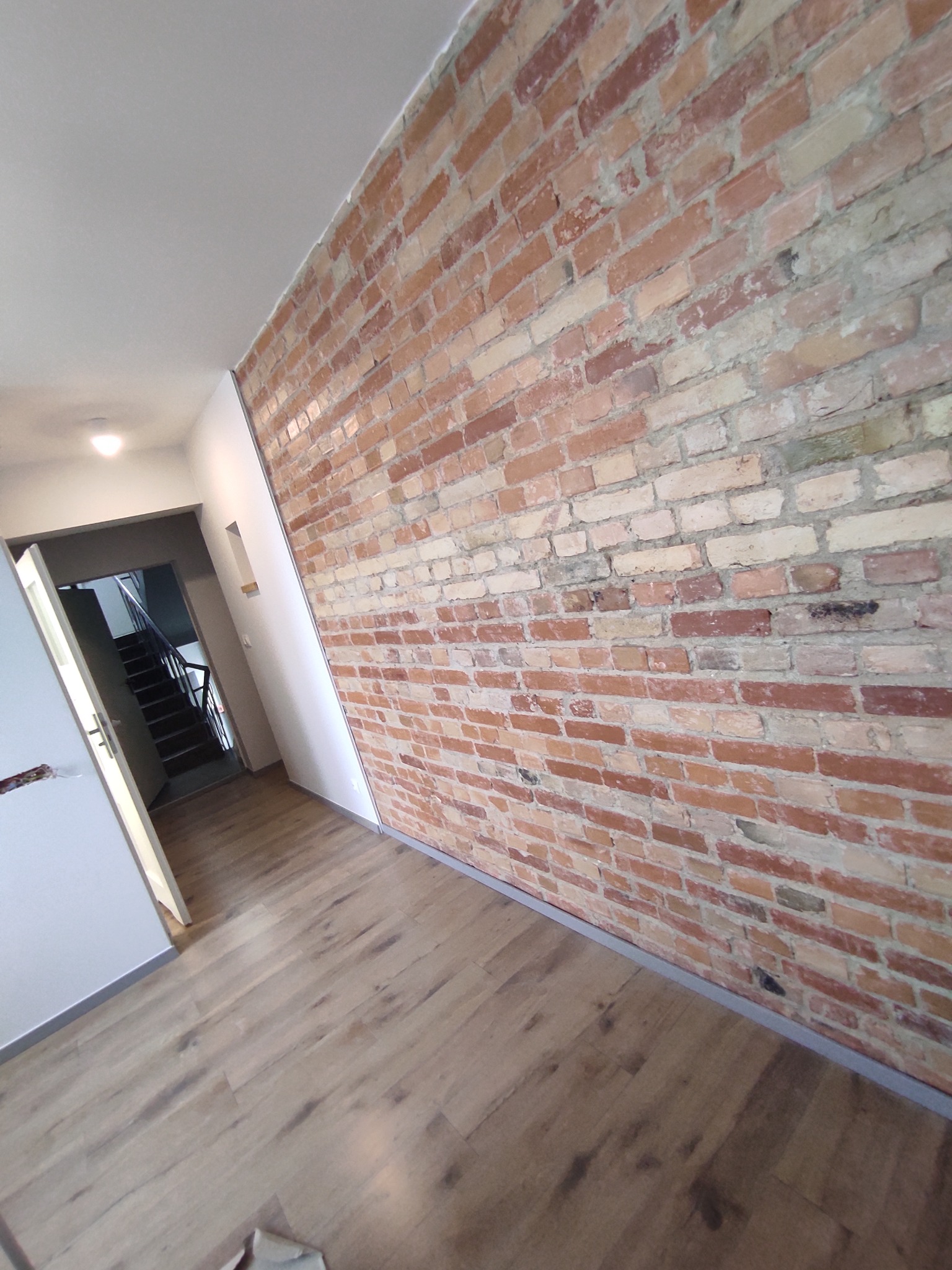 Wnętrze z ceglaną ścianą, drewnianą podłogą i widokiem na schody w tle, ukazujące efekt dekoracyjny i aranżację przestrzeni.