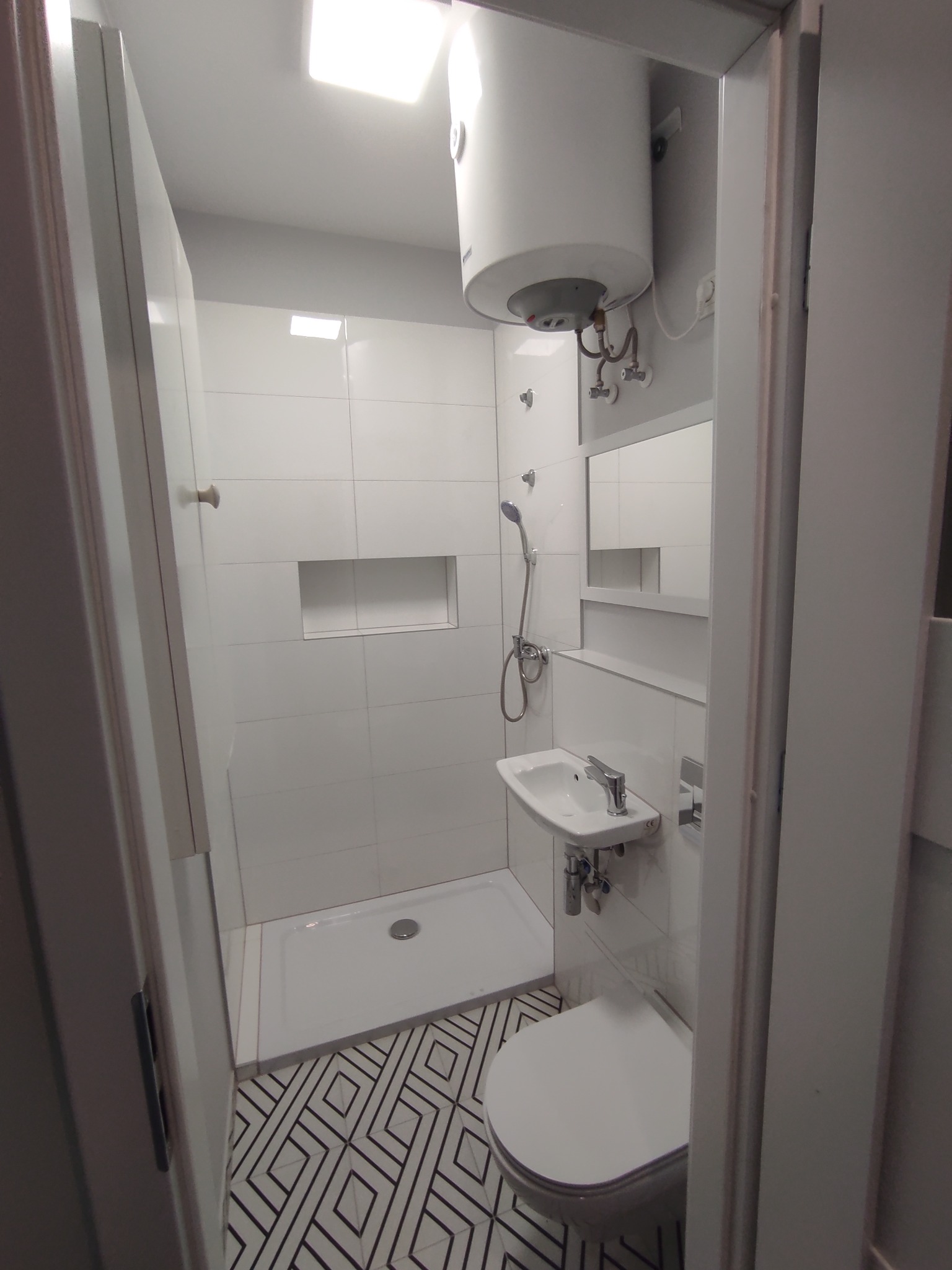 Nowoczesna łazienka z kabiną prysznicową, umywalką, toaletą i bojlerem, wykończona w bieli z geometrycznym wzorem na podłodze.