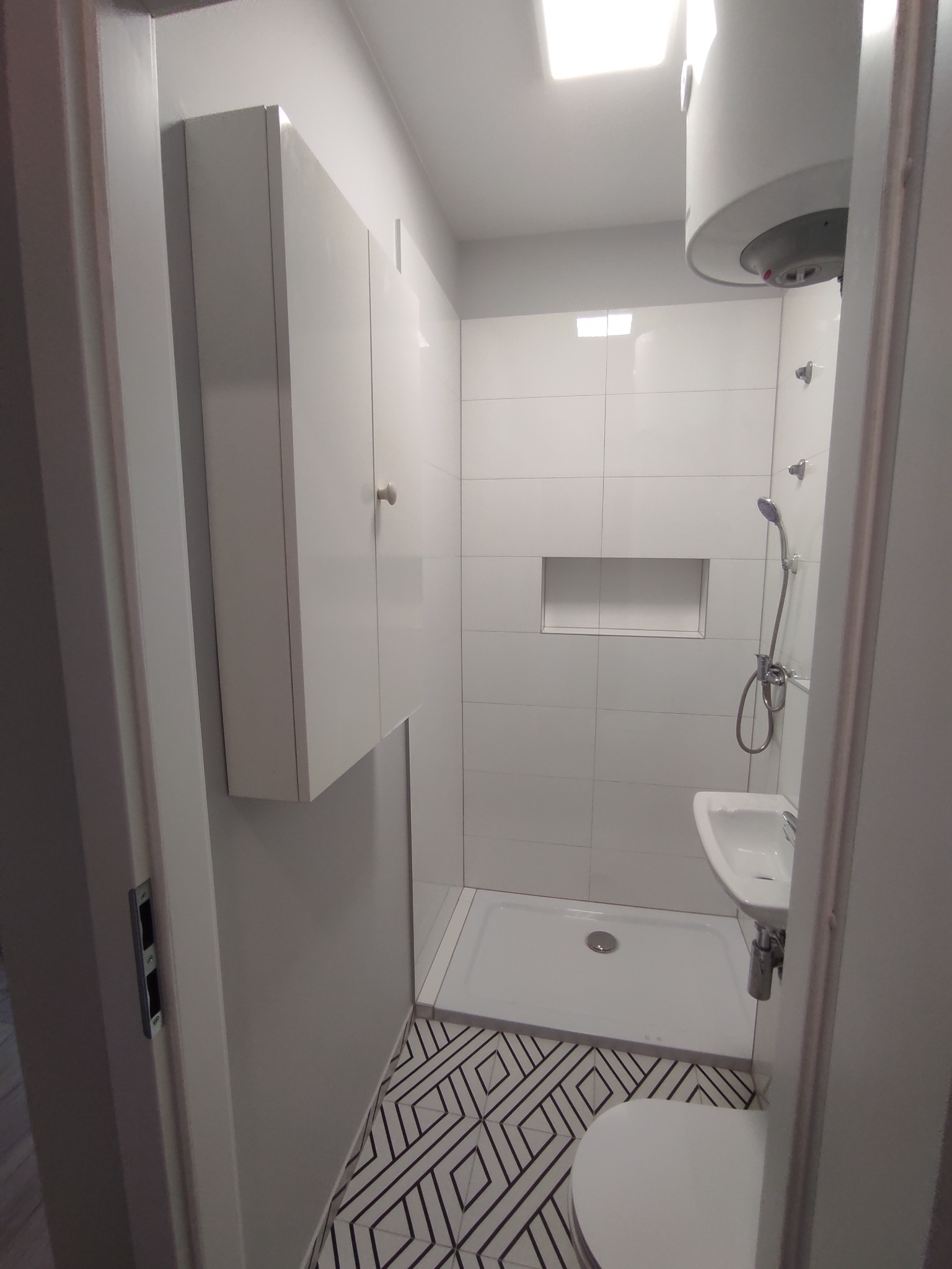 Nowoczesna łazienka w odcieniach bieli z geometryczną podłogą, kabina prysznicowa z odpływem liniowym, umywalka nablatowa i podgrzewacz wody