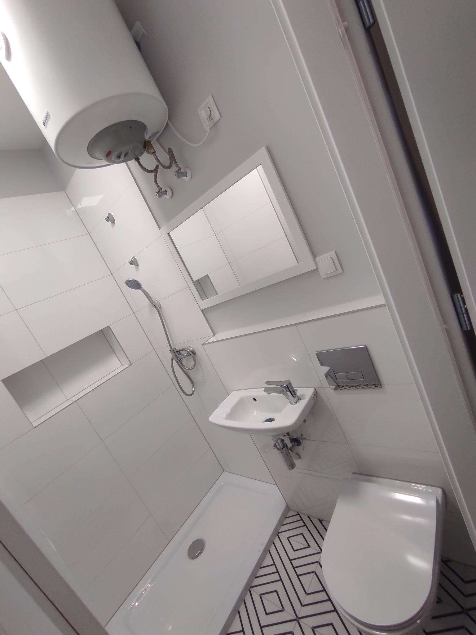 Nowoczesna, minimalistyczna łazienka z białymi płytkami, kabiną prysznicową bez brodzika, podwieszaną umywalką i toaletą, oraz wzorzystą podłogą w czarno-białe romby, z widocznym bojlerem na ścianie.