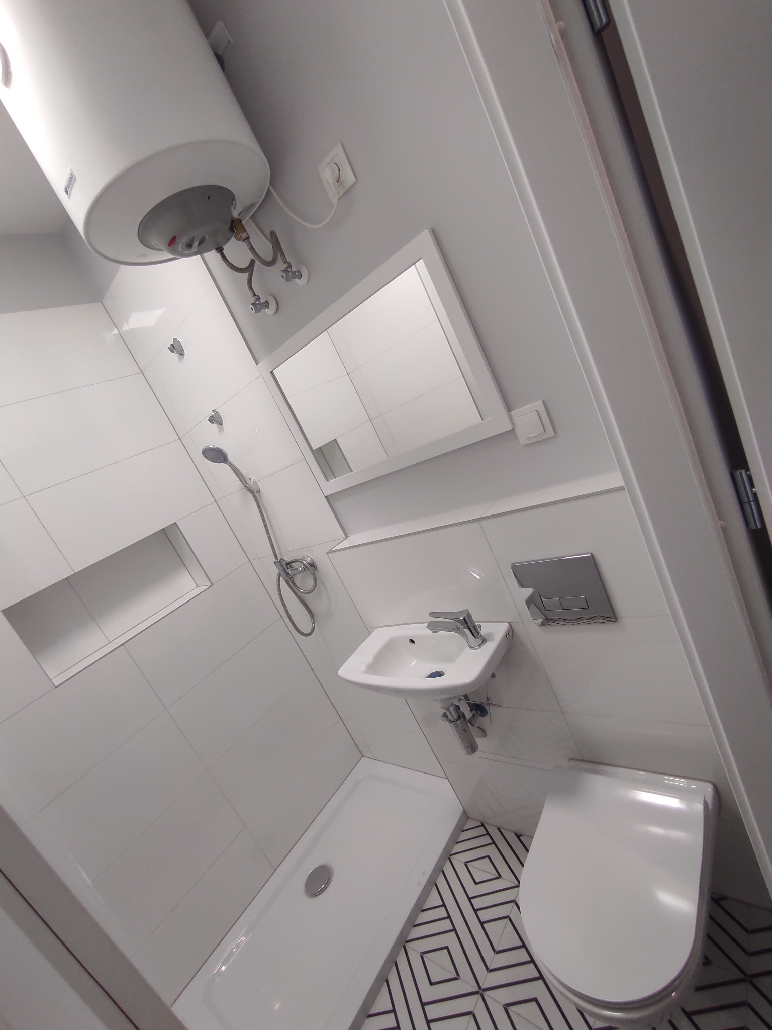 Nowoczesna, biała łazienka po remoncie z wiszącym bojlerem, kabiną prysznicową, umywalką i toaletą na podłodze z geometrycznym wzorem.