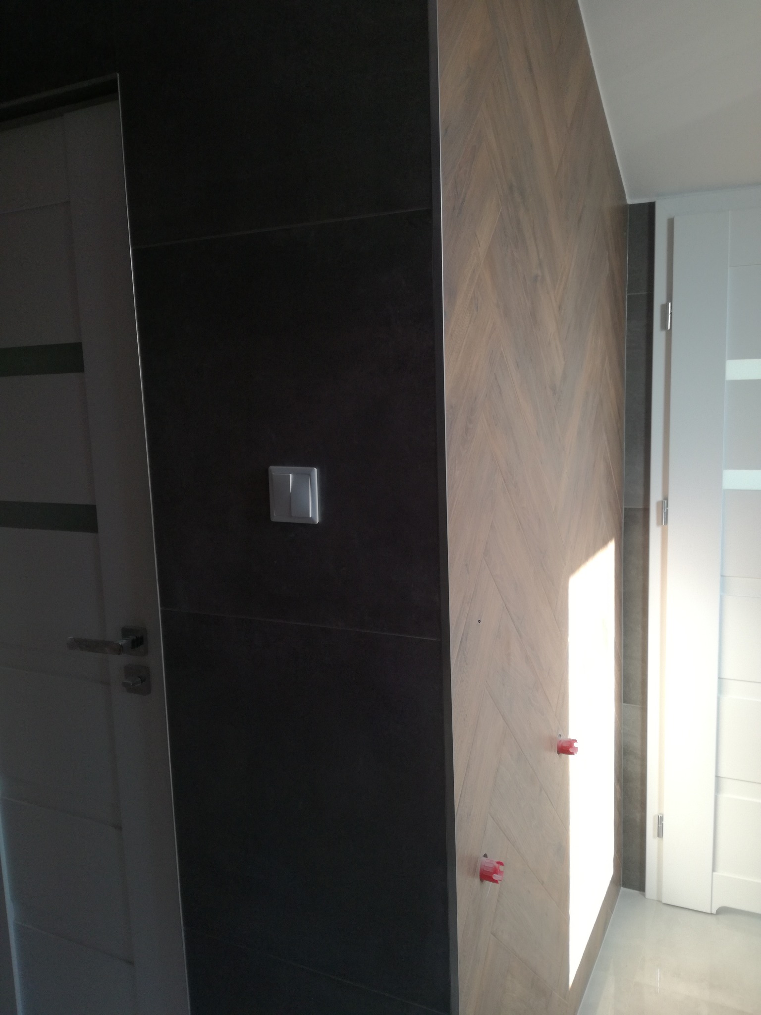 Ściana z ciemnych płytek z włącznikiem światła sąsiaduje z jasną ścianą wyłożoną płytkami w jodełkę, widoczne czerwone krzyżyki dystansowe na płytkach, białe drzwi po lewej i prawej.