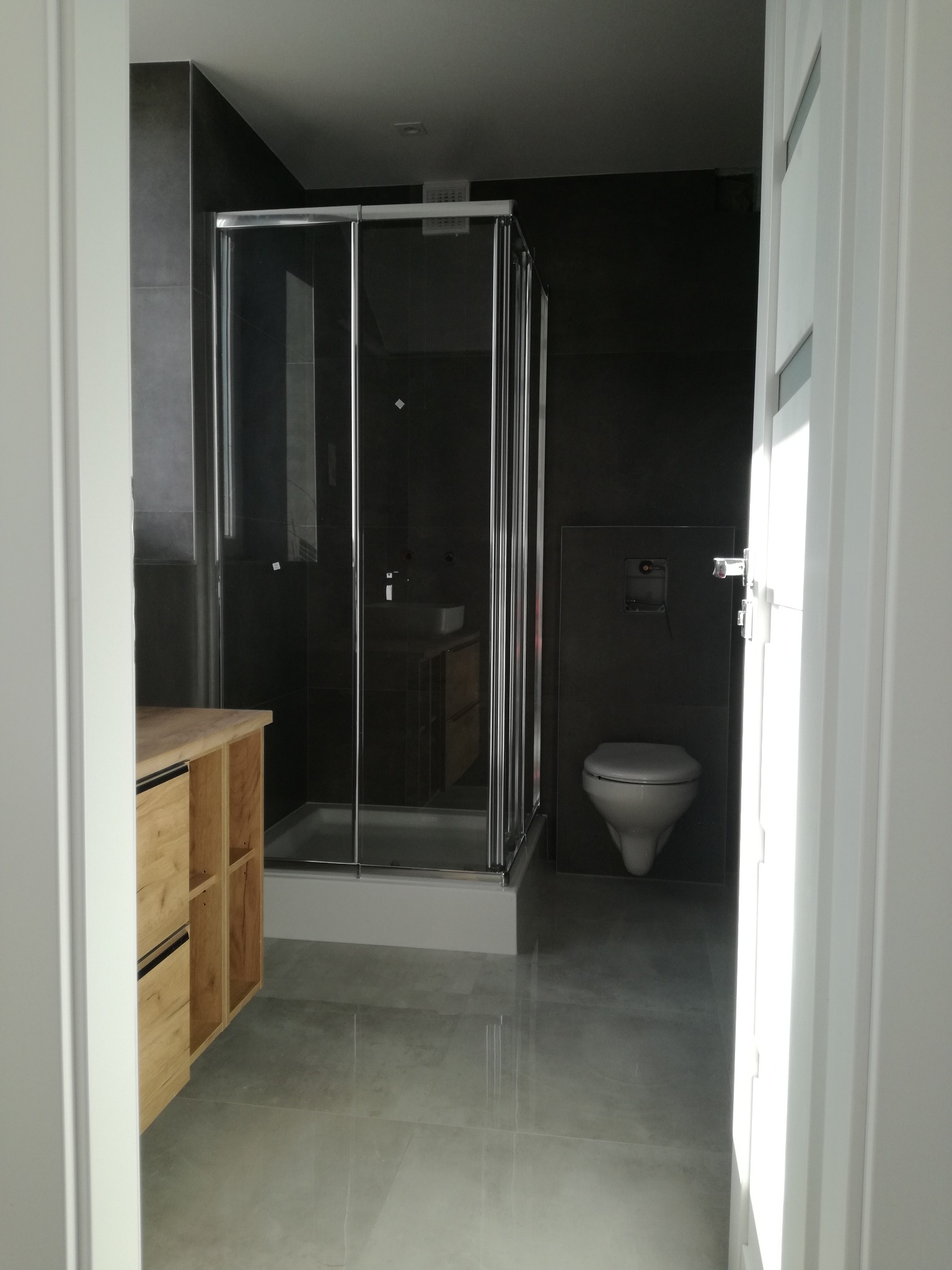 Nowoczesna łazienka z szarymi płytkami, kabiną prysznicową ze składanymi drzwiami, wiszącą toaletą i drewnianą szafką pod umywalką, widziana zza białych drzwi.