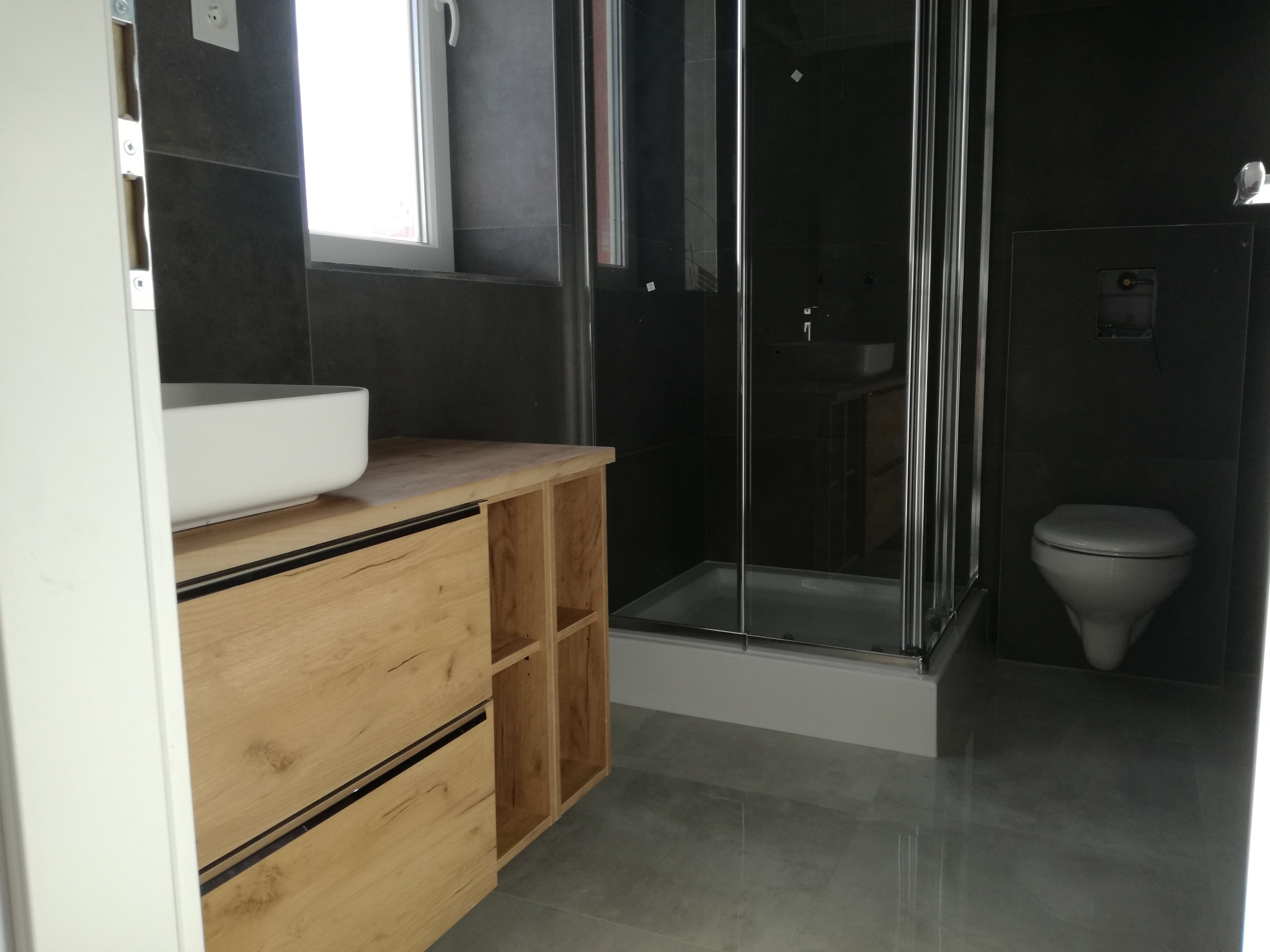 Nowoczesna łazienka z drewnianą szafką pod umywalką, kabiną prysznicową ze srebrnymi ramami i wiszącą toaletą na tle ciemnych płytek.