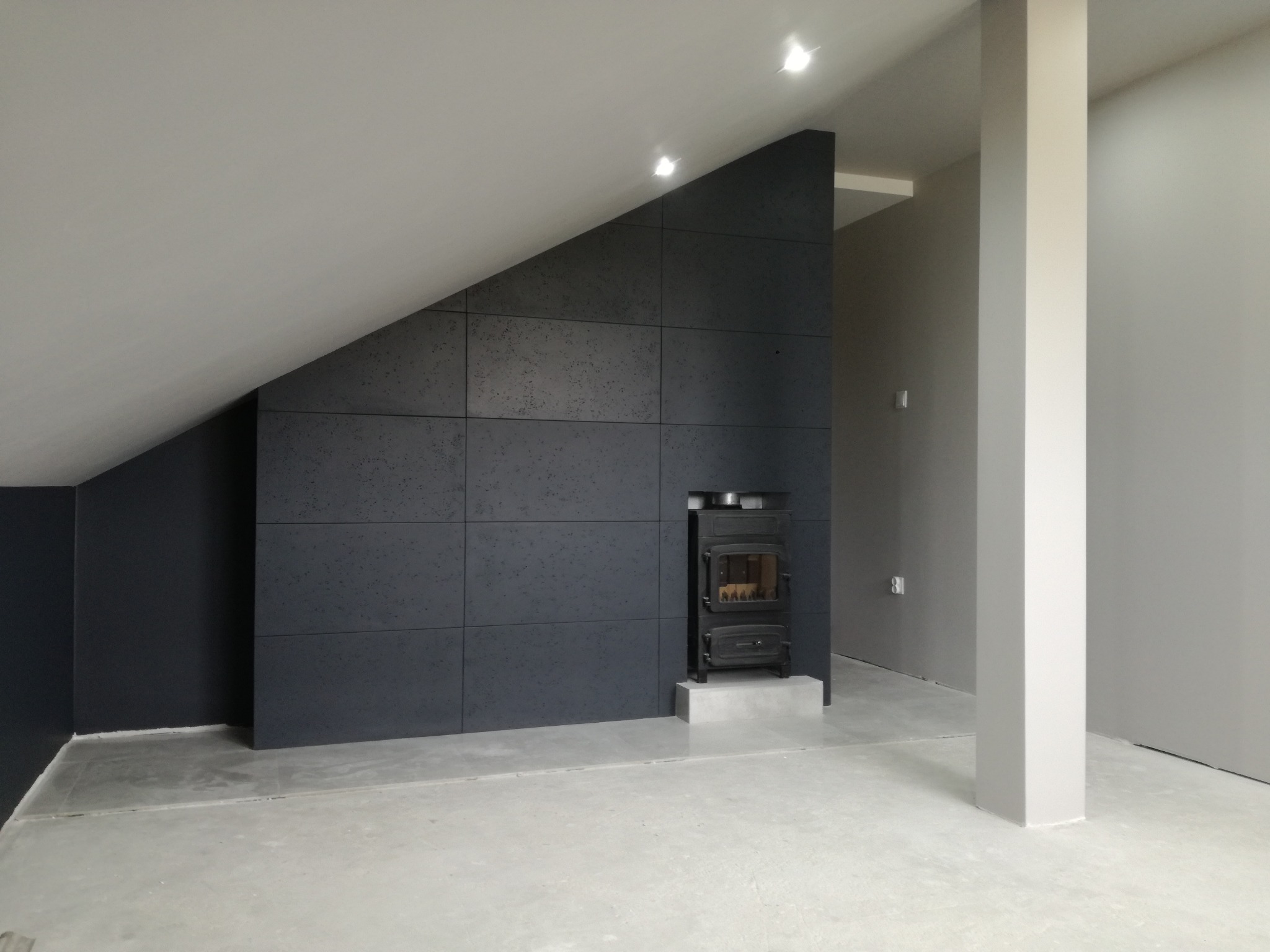 Wnętrze poddasza z kominkiem obudowanym ciemnoszarymi płytami imitującymi beton, szara podłoga i ściany, widoczne oświetlenie punktowe.