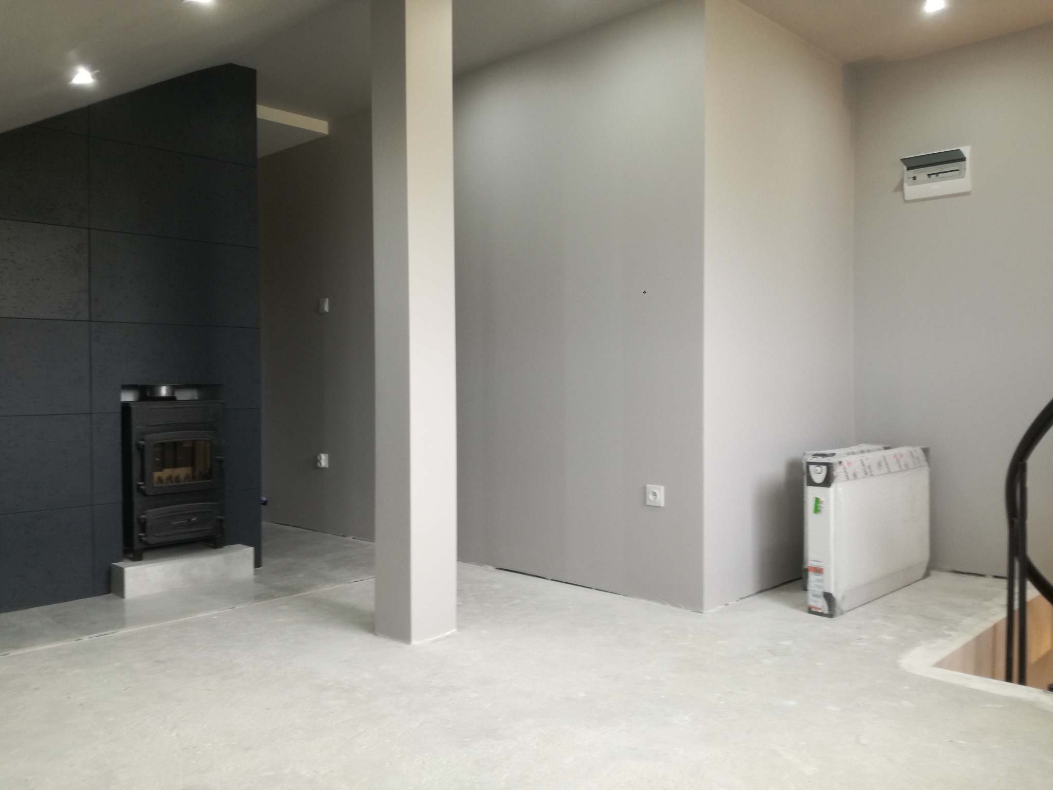 Surowe wnętrze w trakcie wykańczania: czarny kominek z wkładem, szare ściany z instalacją elektryczną, betonowa podłoga i zapakowany grzejnik.