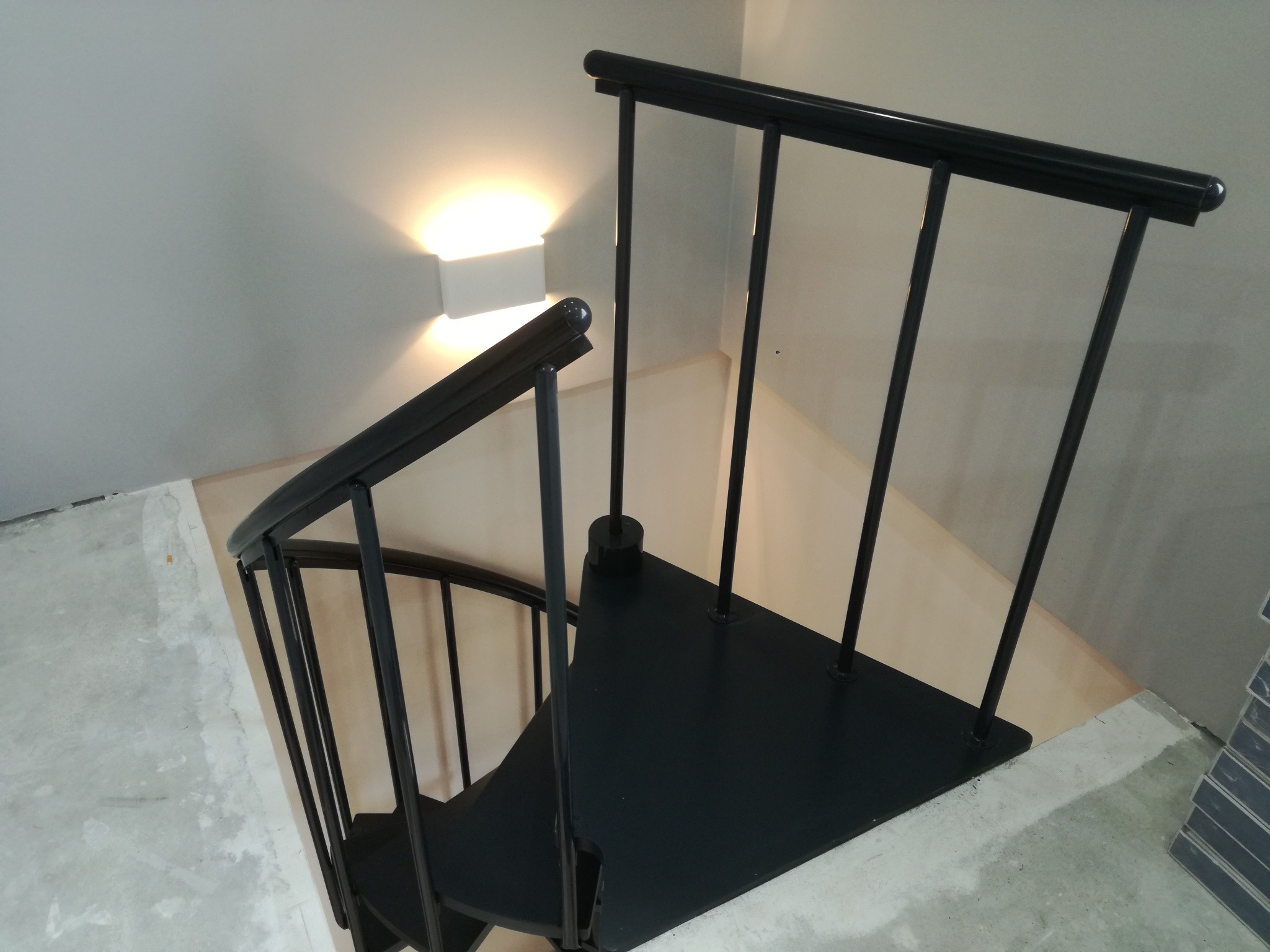 Wnętrze w trakcie remontu: czarne, spiralne schody z metalowymi barierkami, oświetlone kinkietem, na tle surowych, szarych ścian i betonowej podłogi przed wykończeniem.