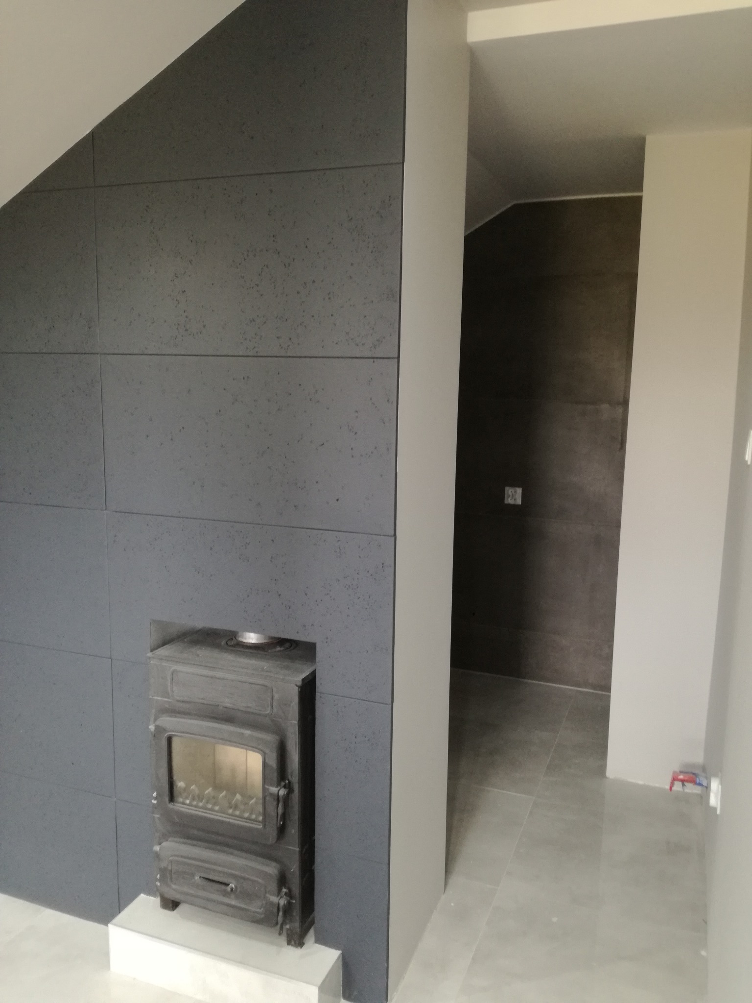 Szara ściana z płyt imitujących beton, skośny sufit i wnęka z zamontowanym żeliwnym piecem wolnostojącym; w tle widoczne przejście do kolejnego pomieszczenia wykończonego w podobnej stylistyce.