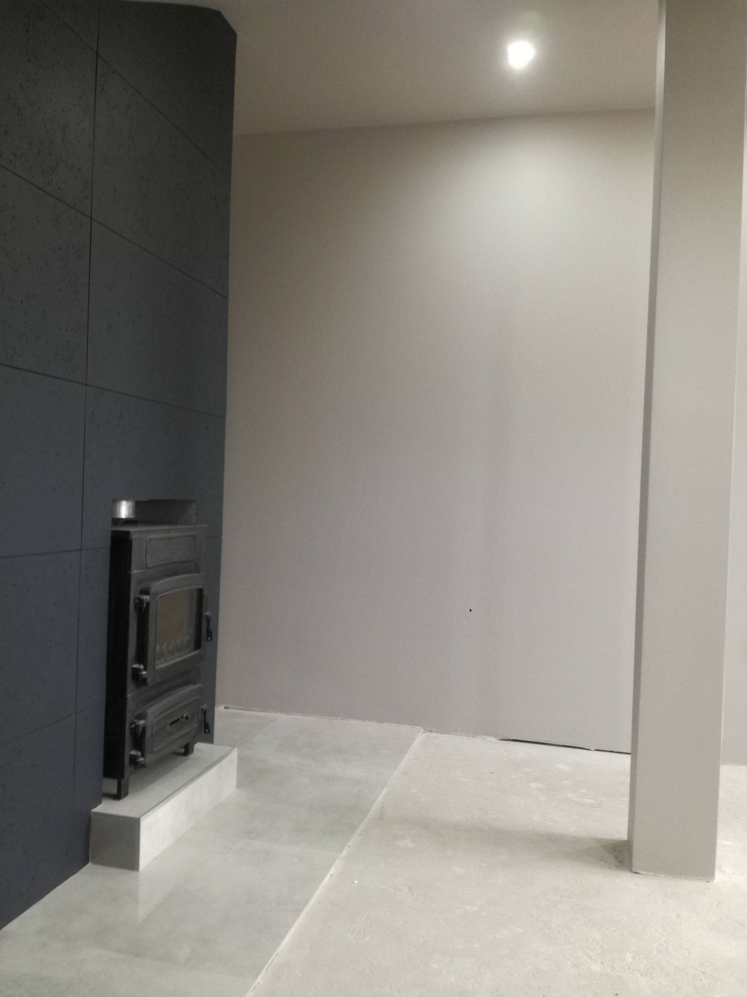 Wnętrze w trakcie wykańczania: szara ściana z płyt, kominek na podeście i jasna podłoga z widocznymi łączeniami płytek.