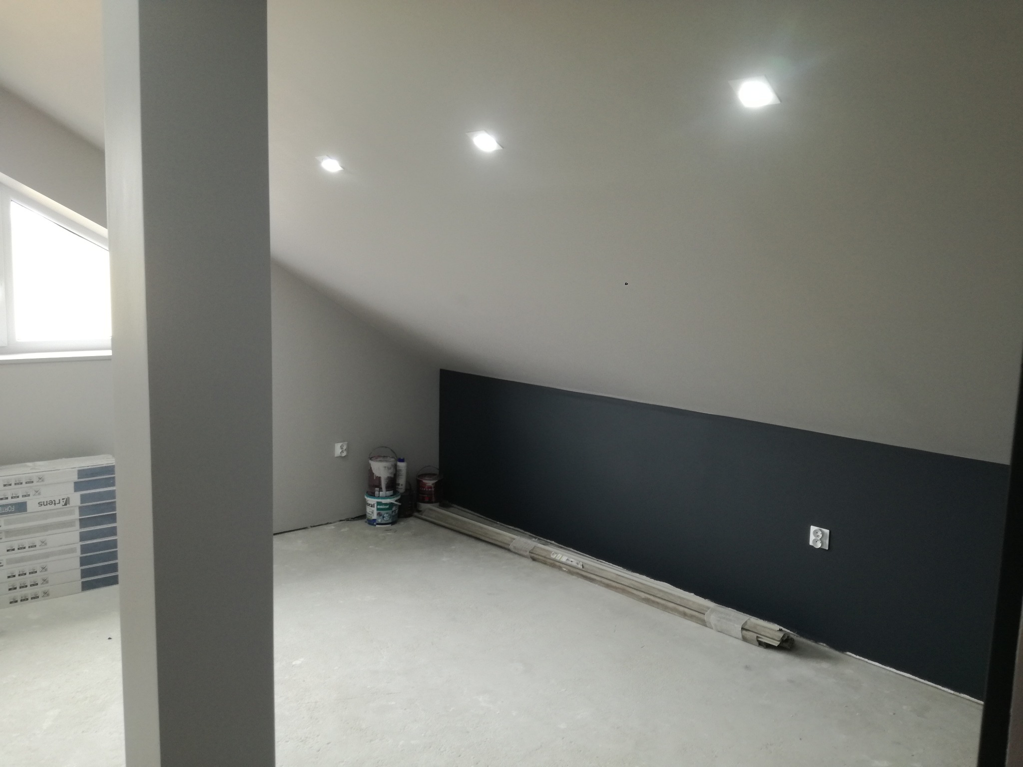 Poddasze w trakcie malowania: ściana pomalowana na ciemny grafit, widoczne puszki z farbą, listwy przypodłogowe i gniazdko elektryczne.