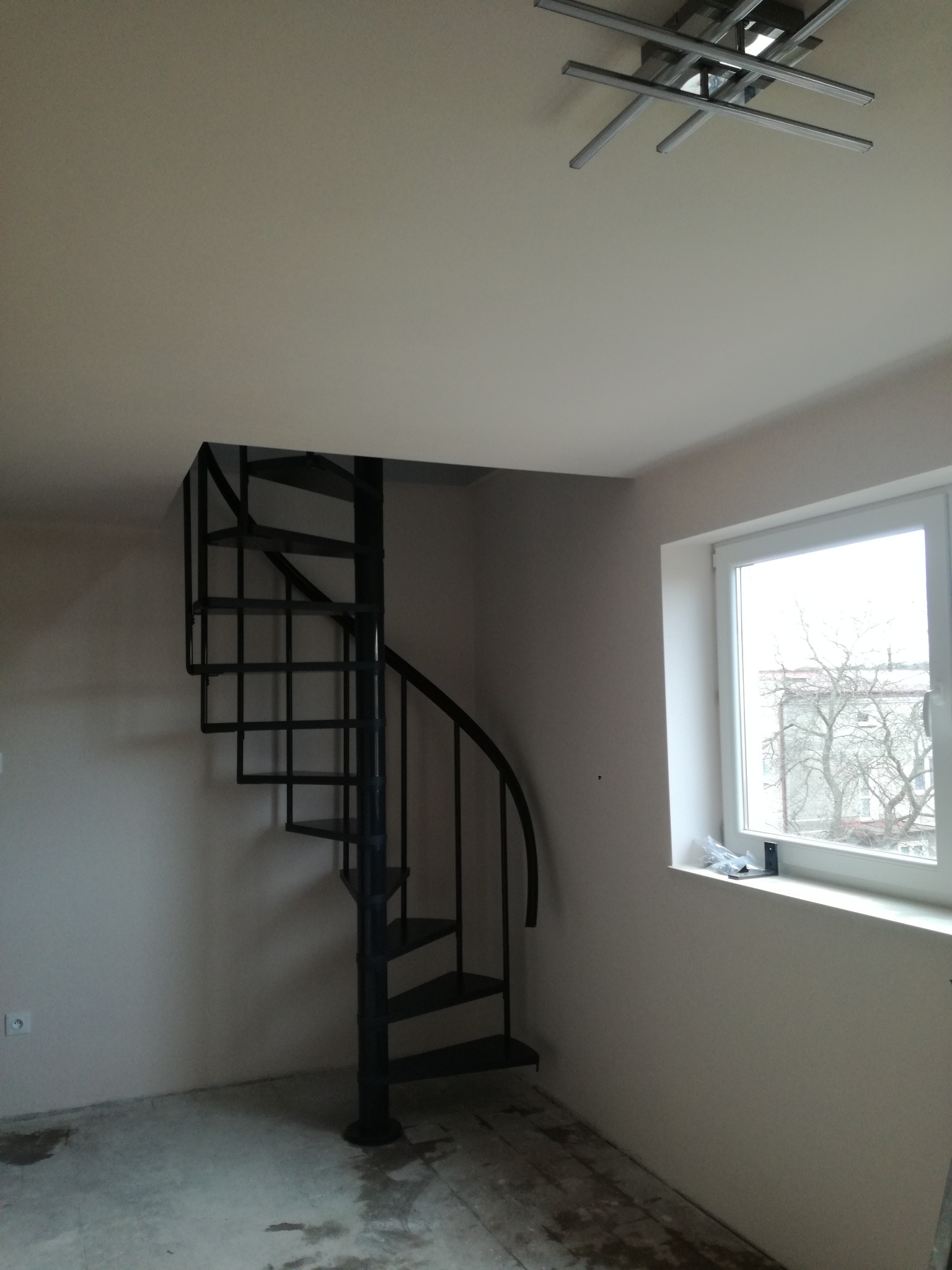 Czarna, spiralna klatka schodowa w jasnym, jeszcze niewykończonym wnętrzu w Świdniku, widoczna z dołu, z oknem po prawej stronie i nowoczesną lampą sufitową.