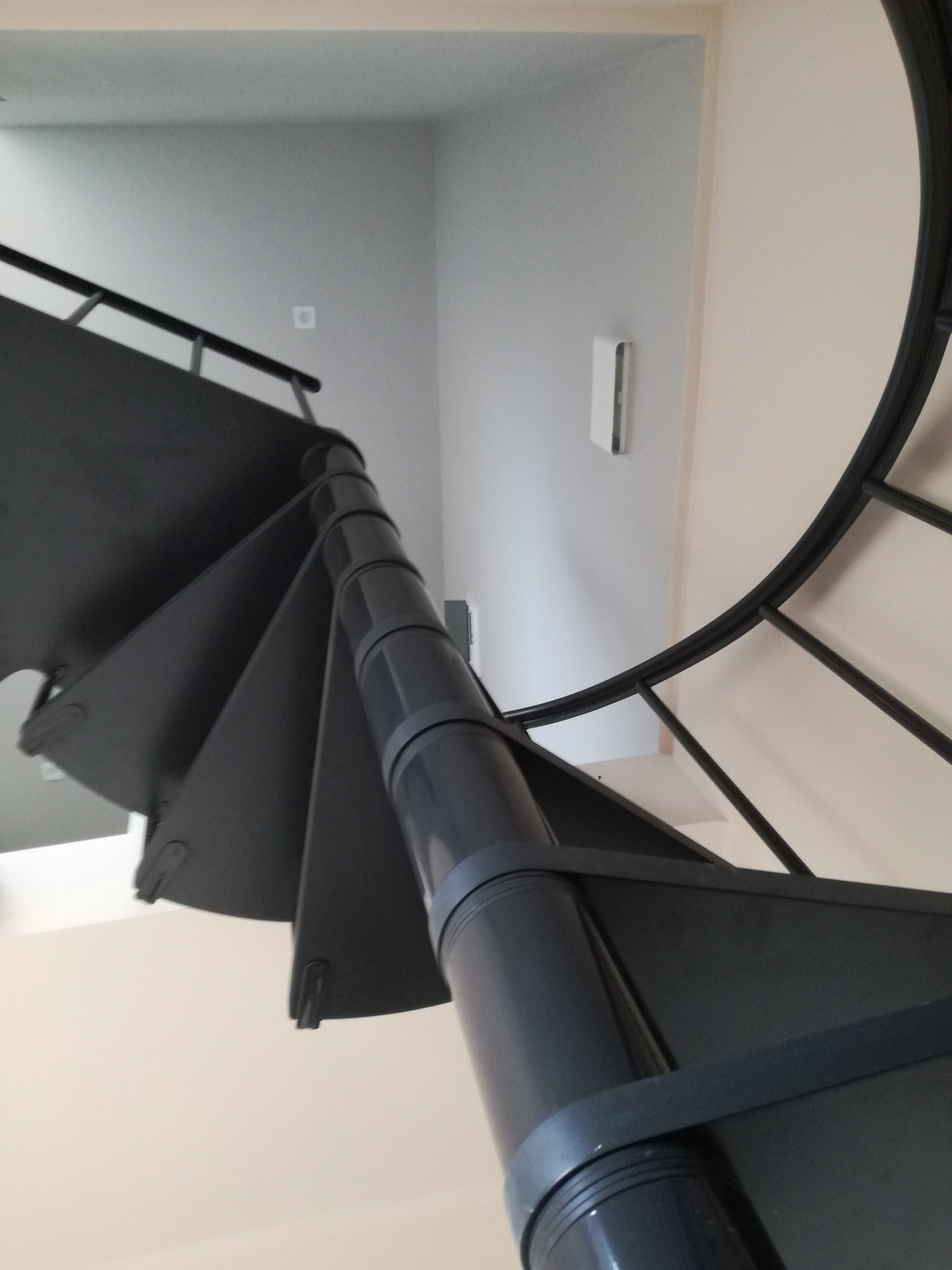 Metalowe schody spiralne wewnątrz jasnego pomieszczenia, widok z dołu ukazujący konstrukcję i detale mocowania stopni.