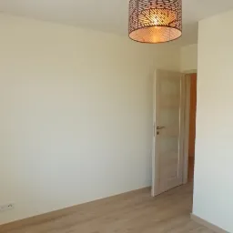 Pusty pokój z jasnymi ścianami, panelami podłogowymi w kolorze drewna i nowoczesną lampą sufitową z ażurowym kloszem, widok na otwarte drzwi do kolejnego pomieszczenia.