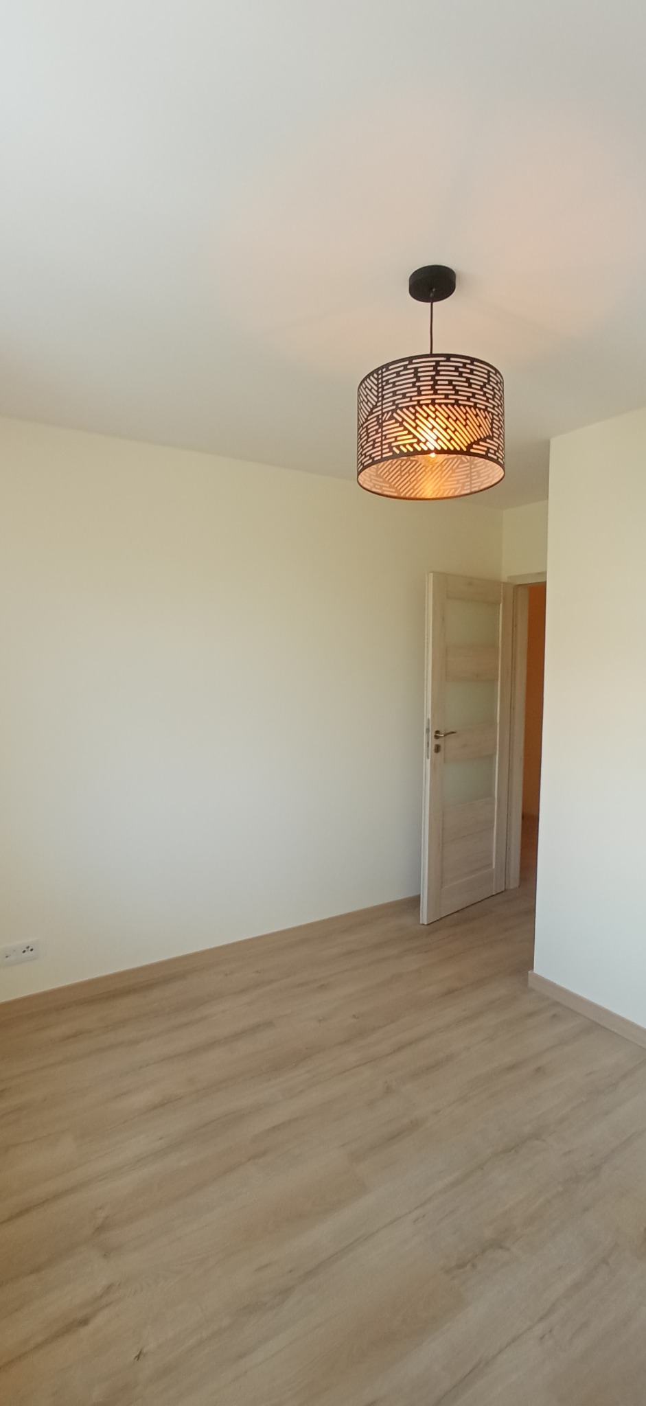 Pusty pokój z jasnymi ścianami, panelami podłogowymi w kolorze drewna i nowoczesną lampą sufitową z ażurowym kloszem, widok na otwarte drzwi do kolejnego pomieszczenia.