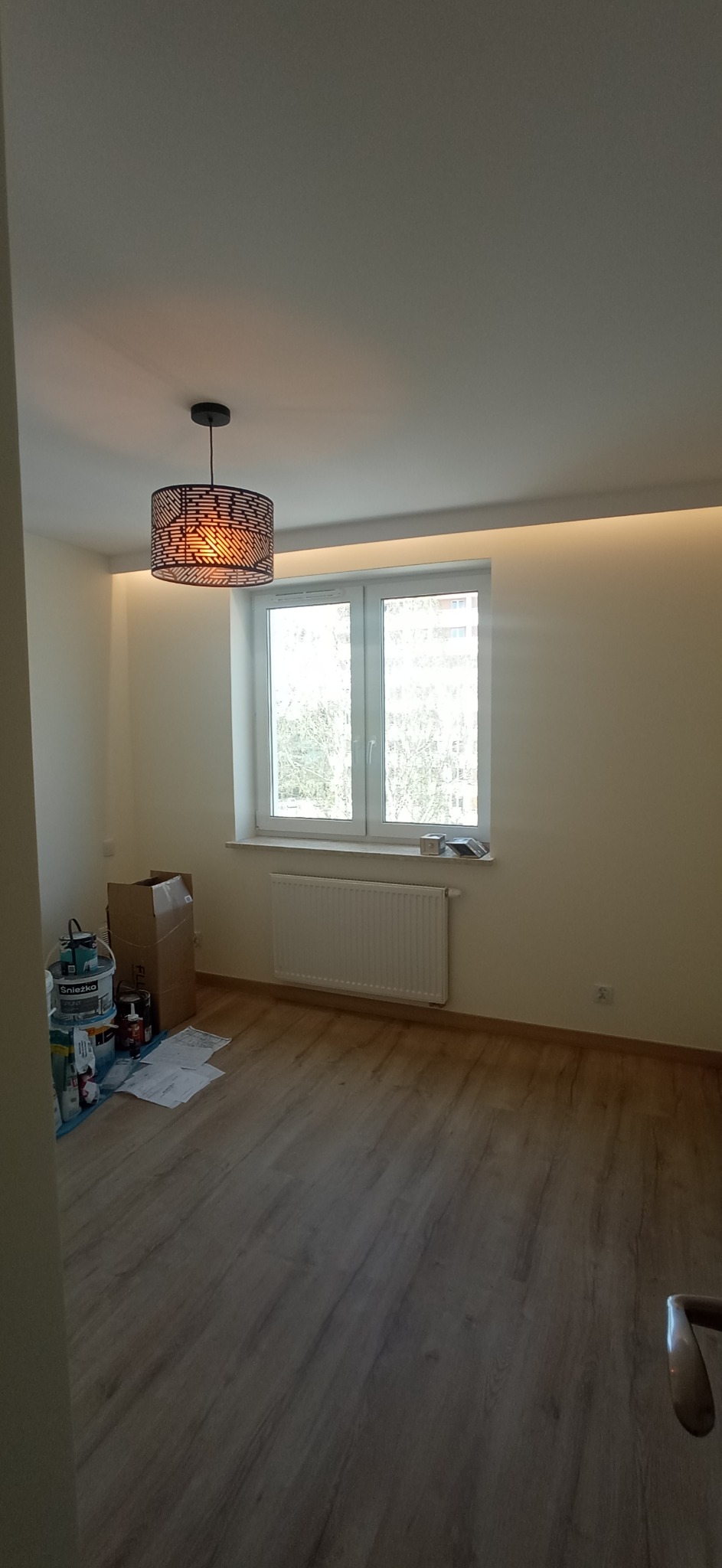 Pokój po malowaniu z panelem podłogowym, widoczne puszki z farbą, lampa wisząca z ażurowym abażurem i oświetlenie LED na suficie.