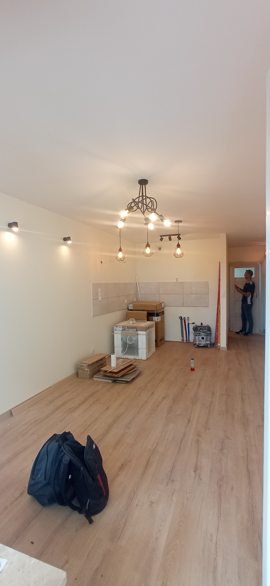 Wnętrze mieszkania w trakcie prac wykończeniowych: położona podłoga z paneli, częściowo wykończona ściana z płytek, wiszące lampy w stylu industrialnym, narzędzia i materiały budowlane, osoba...