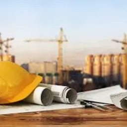 Żółty kask budowlany, zwinięte plany architektoniczne, cyrkiel i ołówek na drewnianym stole. W tle rozmazany widok placu budowy z kilkoma dźwigami.