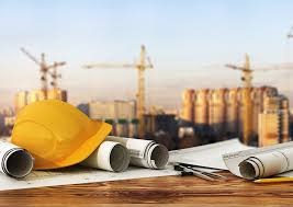 Żółty kask budowlany, zwinięte plany architektoniczne, cyrkiel i ołówek na drewnianym stole. W tle rozmazany widok placu budowy z kilkoma dźwigami.