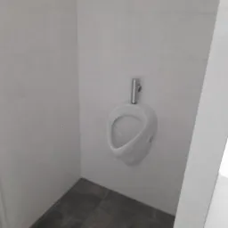 Biały pisuar ścienny z chromowaną rurą doprowadzającą wodę, zamontowany na płytkach w łazience o szarych płytkach podłogowych i jasnych ścianach.