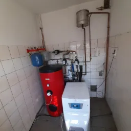 Kompaktowa kotłownia w małym pomieszczeniu z wykafelkowanymi ścianami, widoczny czerwony zasobnik ciepłej wody, biały piec kondensacyjny, zbiorniki wyrównawcze i orurowanie miedziane.