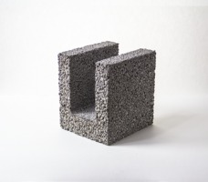 Szary blok z porowatego materiału w kształcie litery U, na białym tle, oświetlony z góry, widoczne drobne nierówności powierzchni.