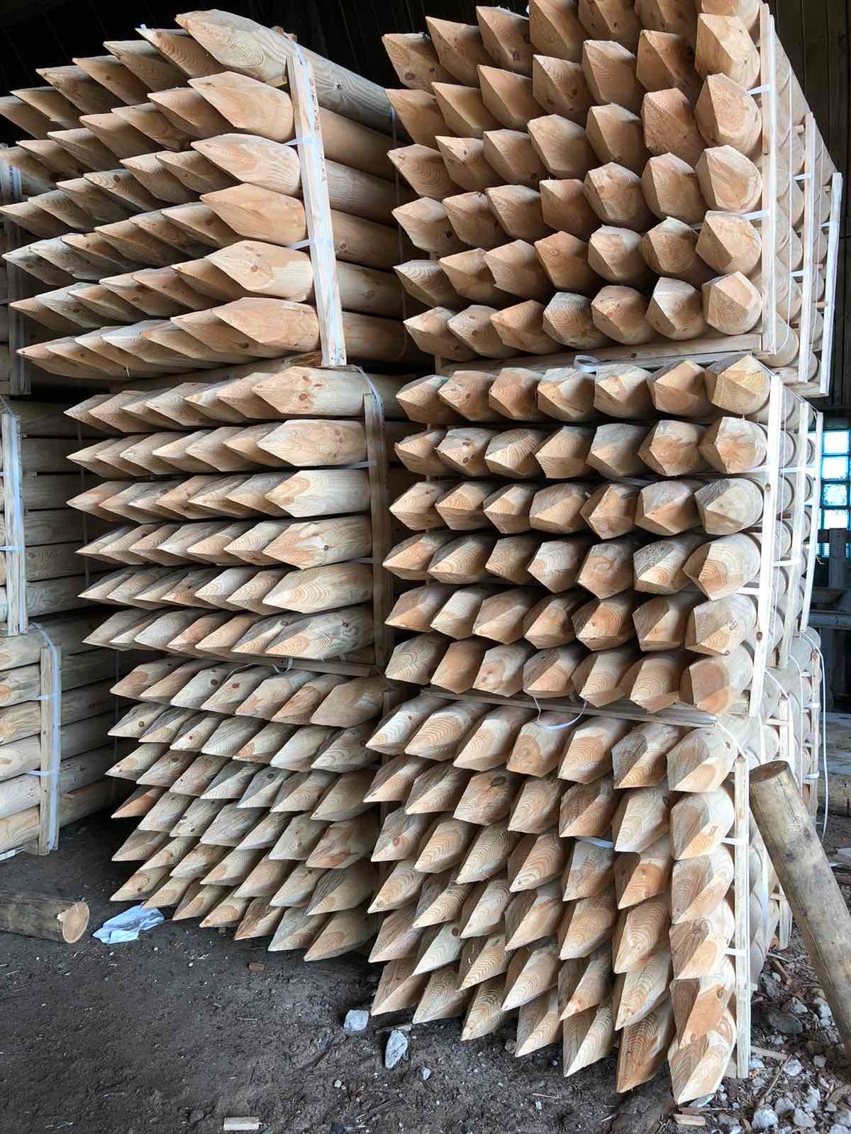 Stosy drewnianych, zaostrzonych pali budowlanych ułożonych w paletach, widoczne słoje drewna i jasne barwy, w tle fragment okna.