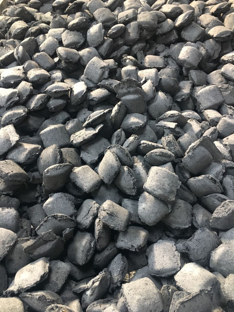 Stos szaro-czarnych kostek opału z widocznymi drobnymi pęknięciami na powierzchni, ułożonych nieregularnie, oświetlonych naturalnym światłem.