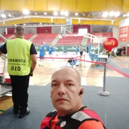 Autoportret ratownika medycznego w pomarańczowej kamizelce z naszywką medyczną na tle hali sportowej z trybunami i grającymi koszykarzami, w tle widoczny steward w żółtej kamizelce.