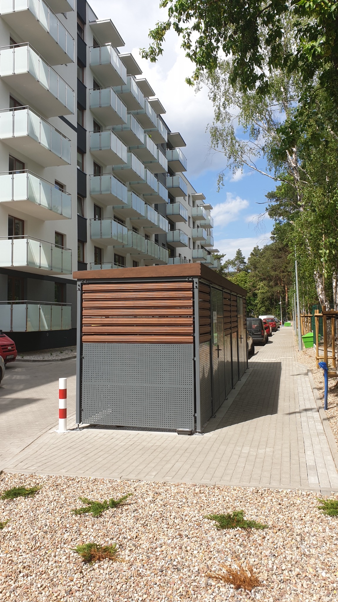 Nowoczesna, metalowa konstrukcja z drewnianymi panelami, prawdopodobnie śmietnik lub schowek, na tle bloku mieszkalnego z balkonami i chodnika z drzewami.