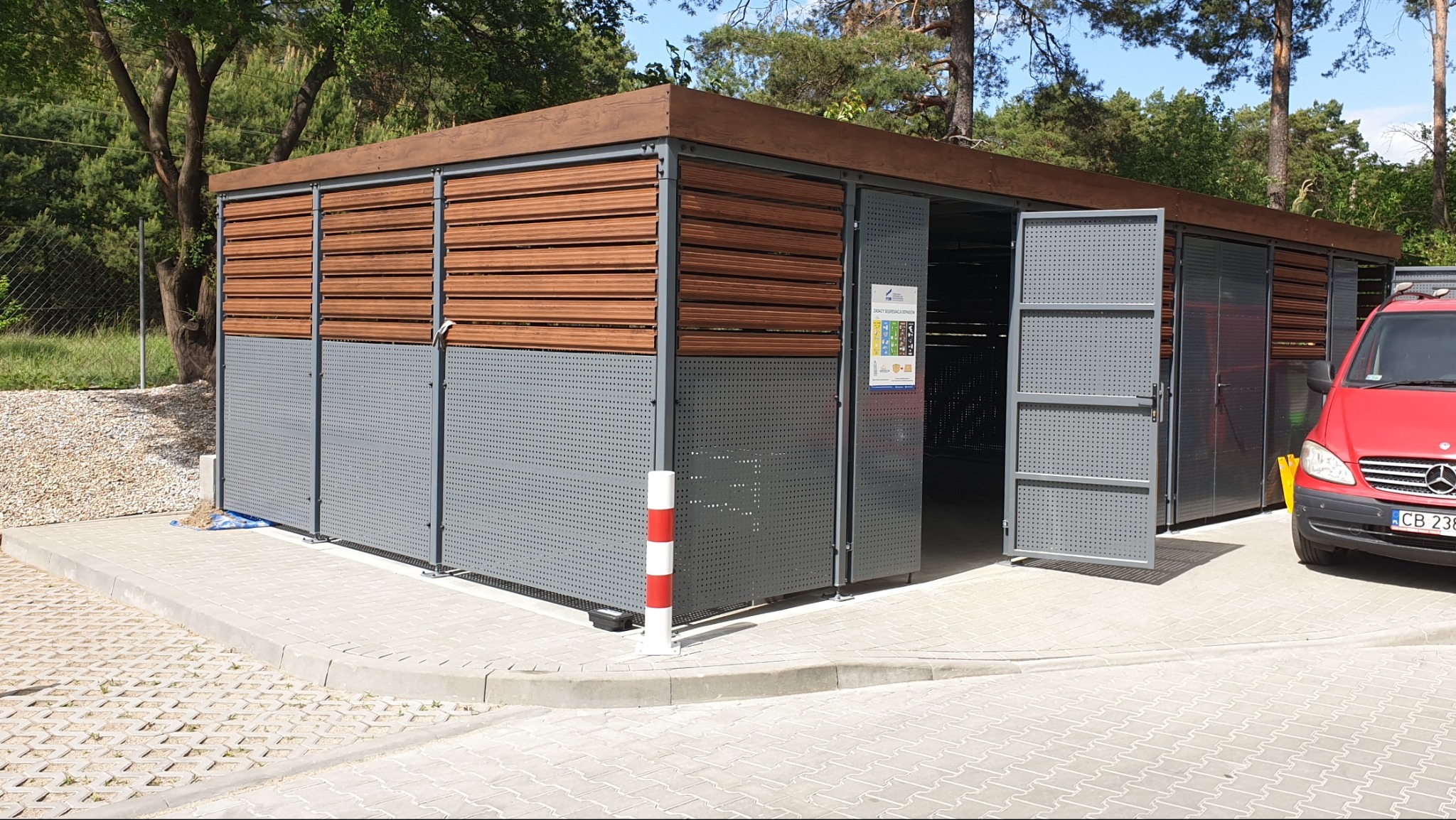 Zewnętrzna wiata śmietnikowa z szarej perforowanej blachy i poziomych drewnianych paneli na górze, z otwartymi drzwiami i czerwonym vanem w tle.