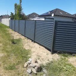 Nowoczesne, szare ogrodzenie panelowe z poziomymi lamelami na tle trawnika i domów jednorodzinnych w słoneczny dzień.