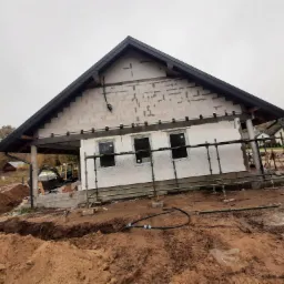 Dom w stanie surowym zamkniętym z widocznym ociepleniem ścian, rusztowaniami i wykopem pod fundamenty na gliniastej działce w pochmurny dzień.