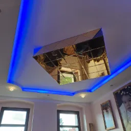 Sufit z dekoracyjnym lustrzanym panelem w kształcie rombu, podświetlony niebieskim światłem LED, widok z dołu na dwa okna z czarnymi ramami i fragmenty mebli w stylu barokowym.