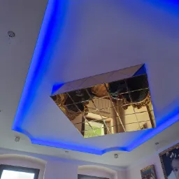 Sufit podwieszany z niebieskim oświetleniem LED i lustrzaną dekoracją w złotej ramie, odbijającą fragment pokoju.