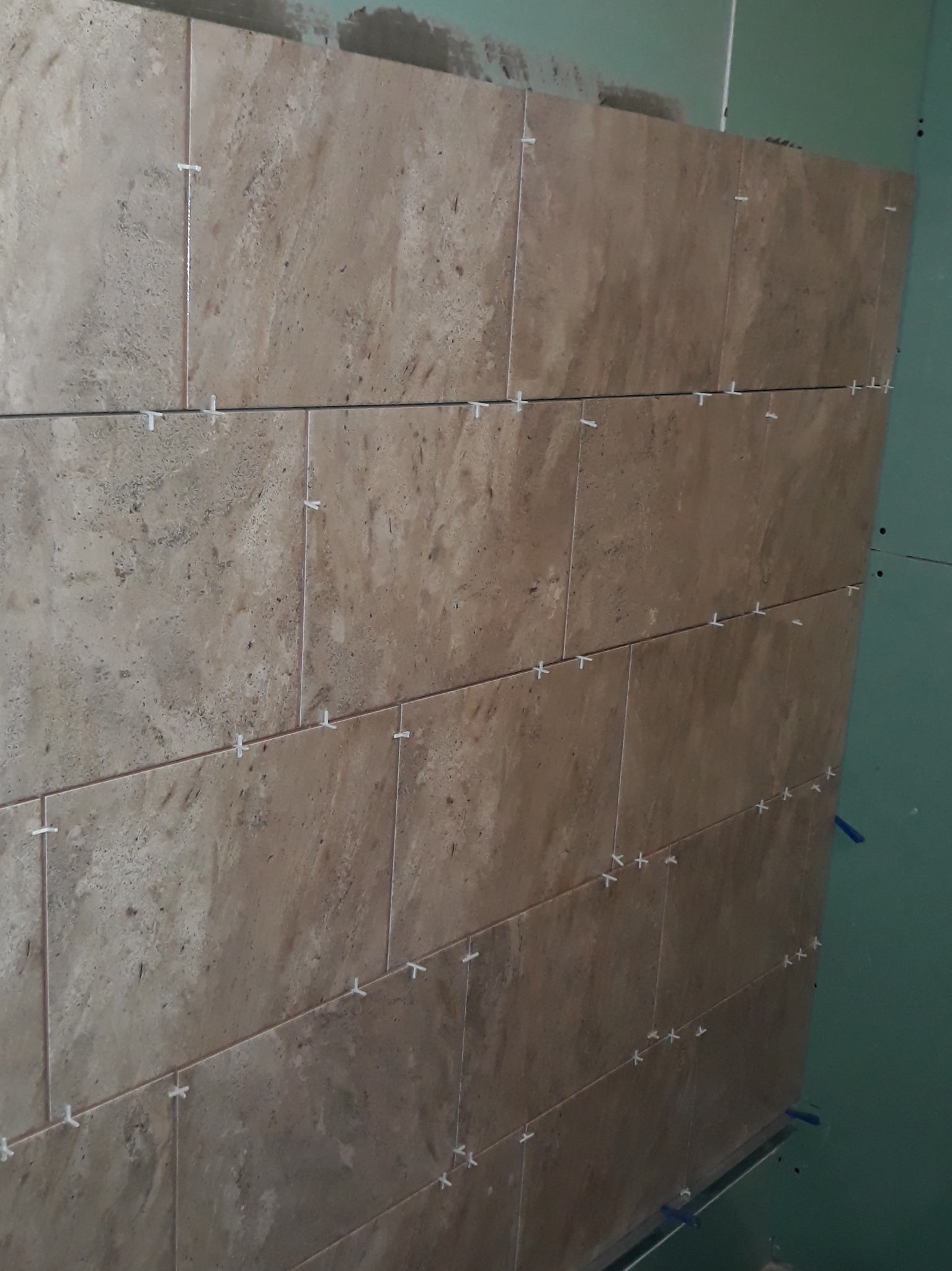 Układanie beżowych płytek imitujących kamień na ścianie z płyt kartonowo-gipsowych, widoczne krzyżyki dystansowe i fragment niezabudowanej przestrzeni na górze.