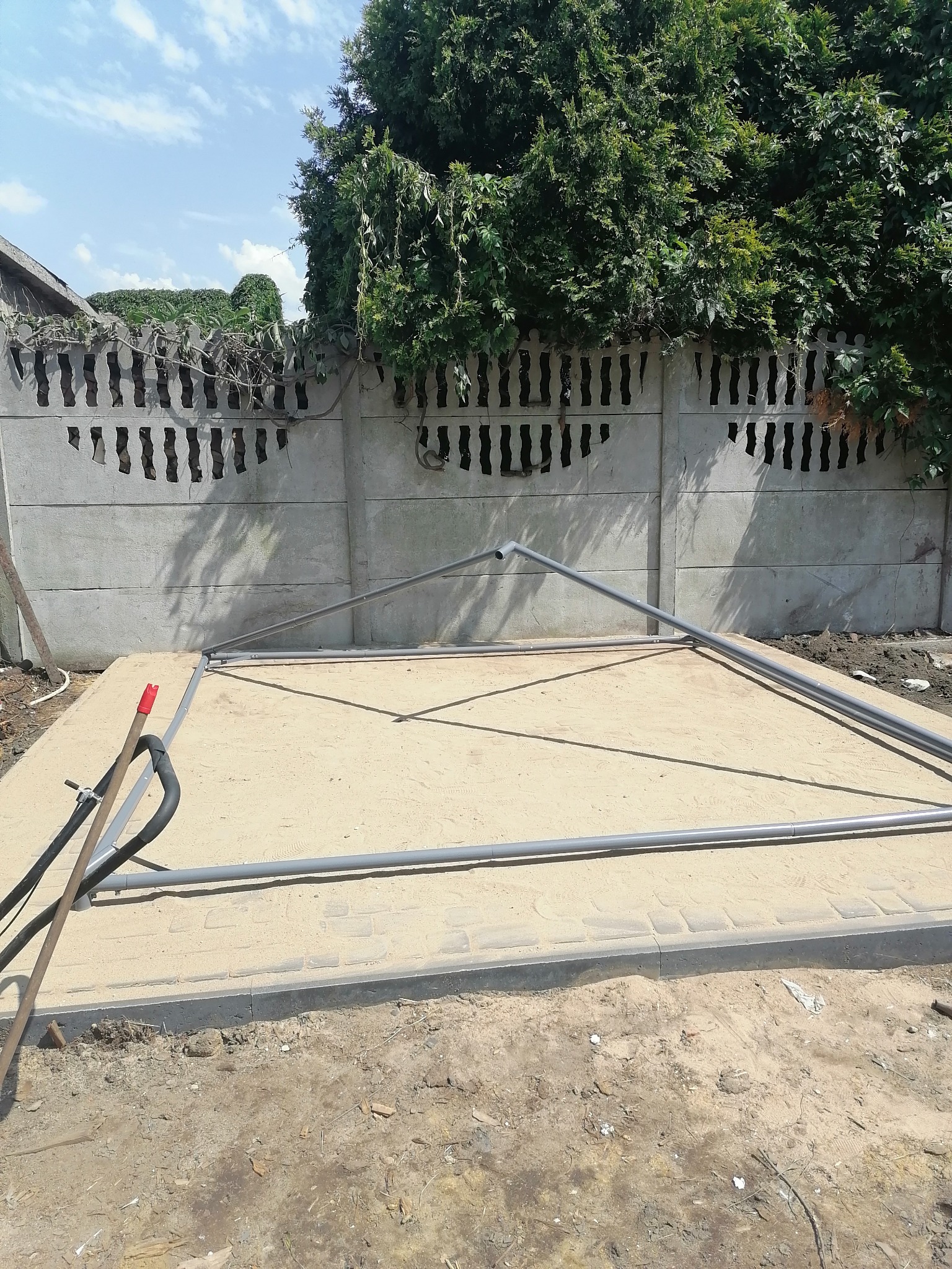 Przygotowanie podłoża pod konstrukcję z rur PCV, widoczny fundament z kostki brukowej, betonowy płot z dekoracyjnymi otworami i zieleń w tle.