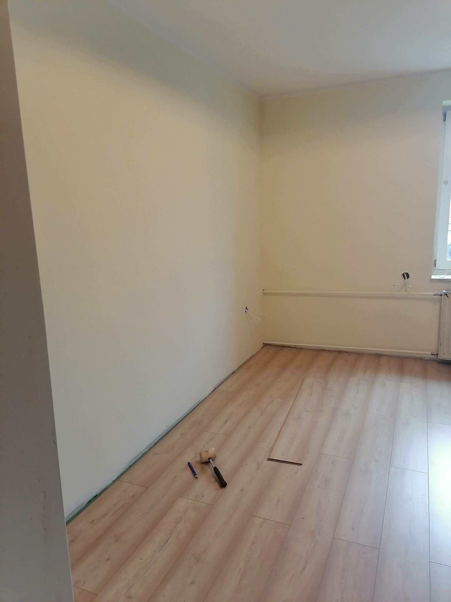 Pokój w trakcie układania paneli podłogowych; widoczny gumowy młotek, ołówek i pojedyncze panele na podłodze, jasne ściany w tle.