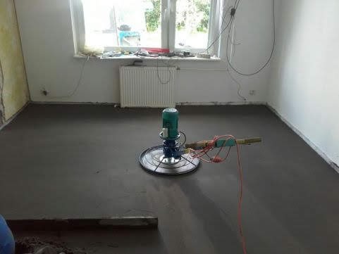 Wygładzanie wylewki jastrychowej w jasnym pomieszczeniu za pomocą zacieraczki mechanicznej; widoczne przewody i niedoskonałości ścian.