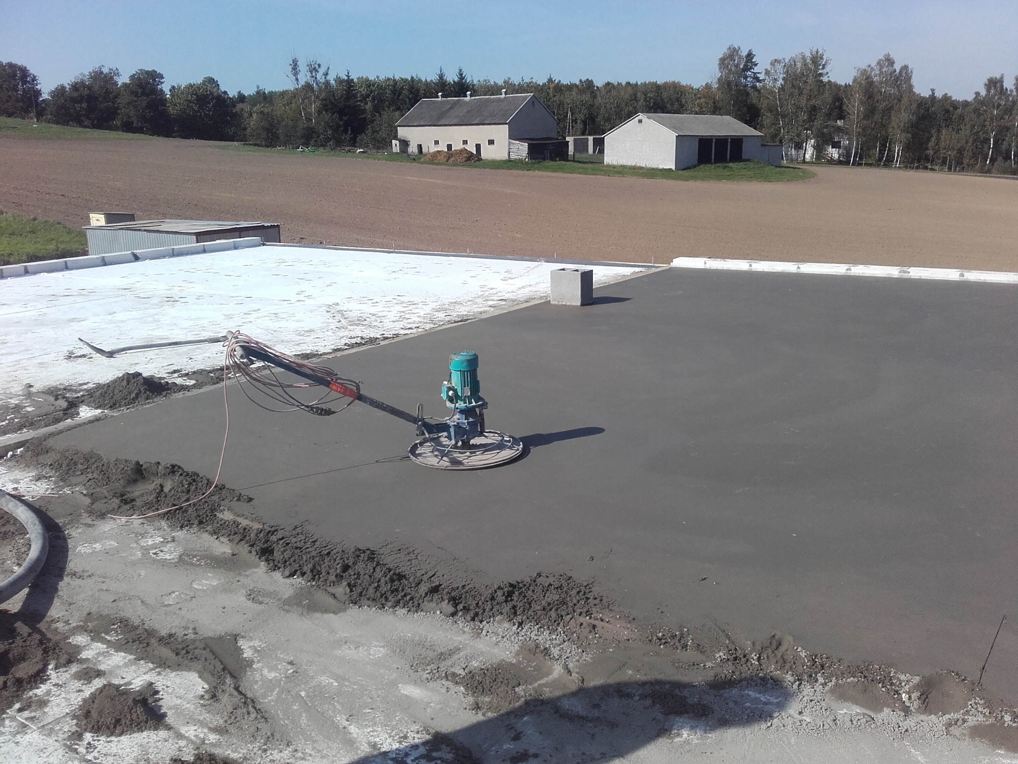 Zacieranie świeżo wylanego jastrychu maszyną zacieraczką na dużej powierzchni, w tle budynki gospodarcze i pole.