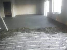 Świeżo wylana, szara posadzka jastrychowa z widocznymi rurami ogrzewania podłogowego w surowym wnętrzu.  Na podłodze widoczne pozostałości zaprawy.