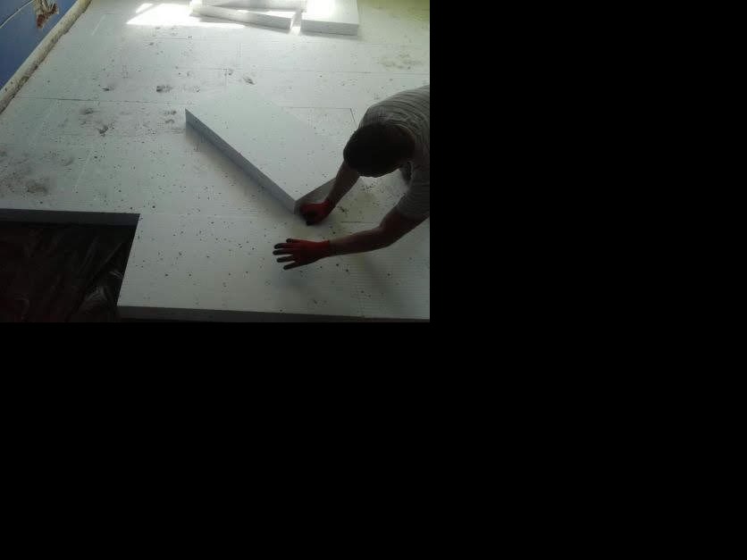 Układanie białych płyt izolacyjnych na podłodze, widoczne ręce w czerwonych rękawicach i fragment ciemnej przestrzeni w podłodze.